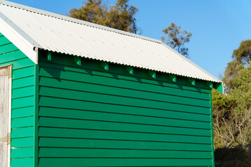 Fototapeta premium Vibrant Green Wooden Beach Hut with Corrugated White Metal Roof Against a Clear Blue Sky on a Sunny Day
