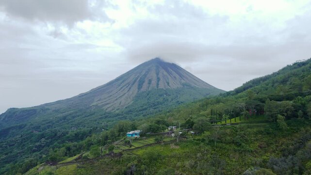 Aerial drone moving forward towards the iconic pyramid-shaped Mount Inerie in Ngada, Flores. Majestic volcano peak surrounded by white clouds.