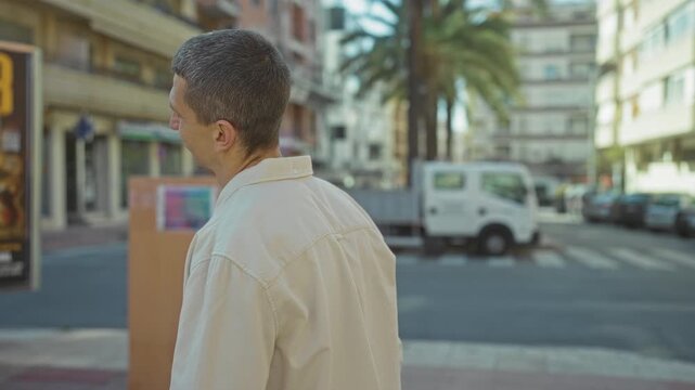 Man standing with crossed arms outdoors on a bustling street with palm trees and urban architecture visible in the background, looking content and thoughtful with short hair.
