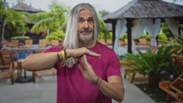Man with long grey hair, hands forming timeout gesture at hotel pool cabana wearing pink shirt and bracelets; calm.
