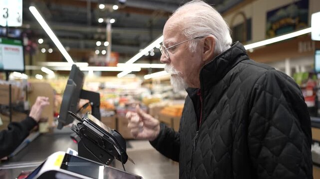 An elderly man faces difficulty at the self checkout while shopping for food at a grocery store in the afternoon.