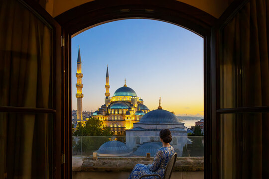 An attractive woman in Middle Eastern dress sits on a balcony at sunset overlooking the New Yeni Mosque and city of Istanbul, Turkey.
