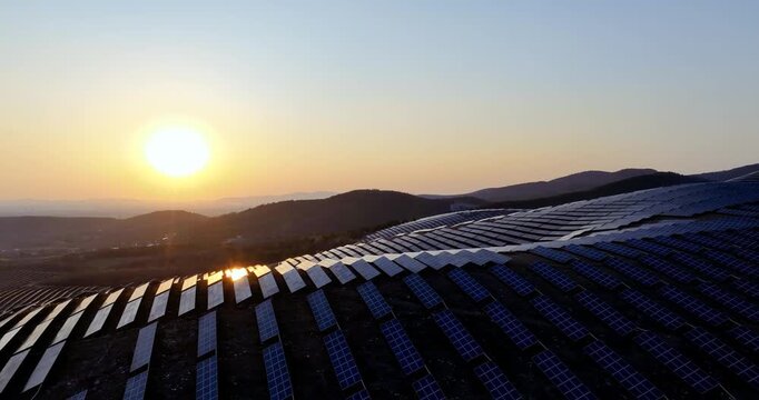 Aerial video of a large solar photovoltaic farm on hills at sunset, with rows of panels reflecting golden light, symbolizing clean energy and sustainable development.