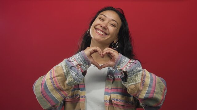 Woman points finger at camera in red studio, wearing patterned jacket and hoop earrings, smiling and forming heart with hands in playful portrait; joy connection.