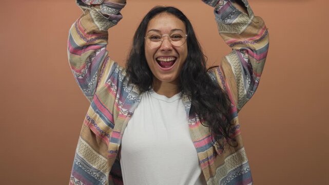 Woman raises arms and smiles broadly wearing glasses and patterned jacket in studio against peach backdrop; joyful celebration.