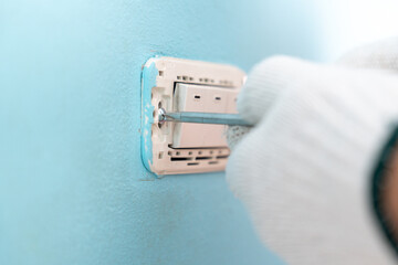 Close-up hand of man wearing cloth gloves and uses a screwdriver to remove a light switch panel for repair.