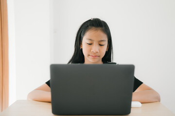 A young girl is sitting at a table, concentrating as she uses a laptop for studying or online learning in a bright, calm home environment.