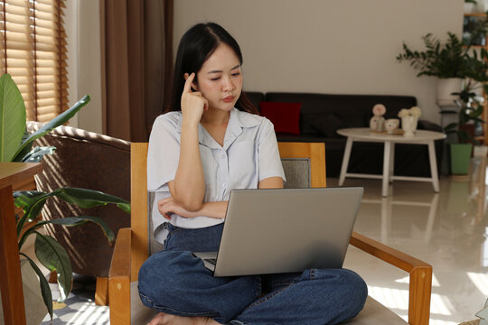 Stressed asian woman holding laptop computer sit alone on chair inside living room feeling tired and headache from work while thinking hard about online job problem