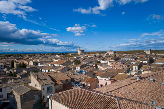 Wide elevated view of textured terracotta rooftops and medieval stone walls in Aigues-Mortes, with the iconic tower and dramatic clouds set against a deep blue spring sky.