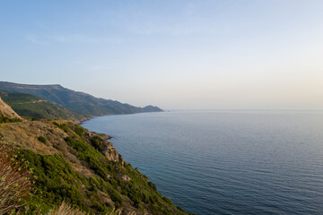 Fototapeta premium A tranquil view of the coastline between Alghero and Bosa, Sardinia, with green hillsides meeting the calm blue Mediterranean Sea under soft, clear evening light.