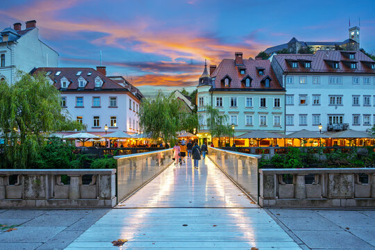 View from the Ribja Brv pedestrian bridge of the illuminated riverbank of sidewalk cafes and shops, with the medieval hilltop castle above, at early evening.