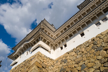 Obraz premium Dramatic upward angle of Himeji Castle's white exterior and layered tiled eaves rising above a textured stone wall, highlighted by sunlight against a partly cloudy sky.