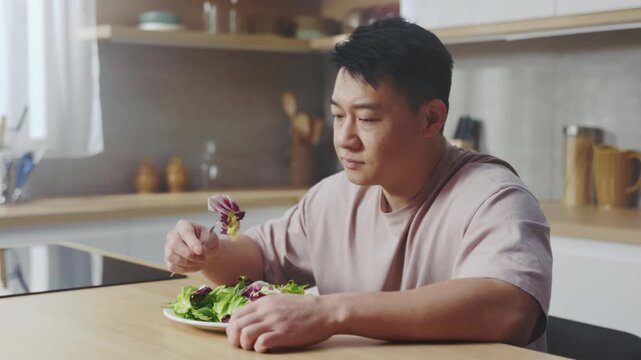 Man carefully tests salad textures at home, Asian man hesitantly evaluates salad in cozy kitchen setting
