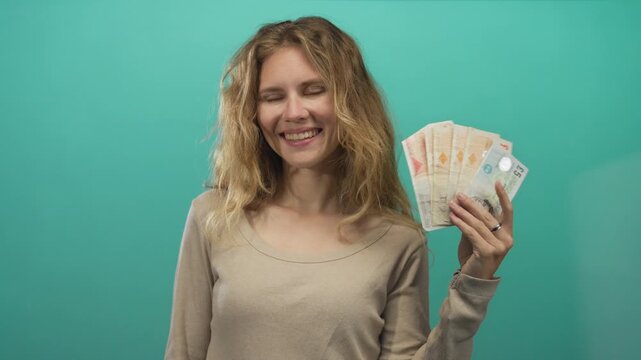 Woman enjoying money while smiling in british campaign shows blonde young investor holding cash and pound notes on studio green background.