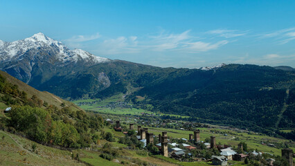 Historic mountain village with medieval towers beneath snowy peaks and bright blue sky.