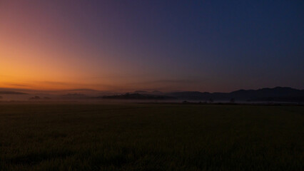 Obraz premium Soft sunrise light over misty fields and distant hills creating a tranquil landscape.