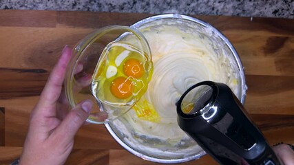 overhead view of person pouring raw eggs into cheesecake batter while mixing with an electric hand mixer