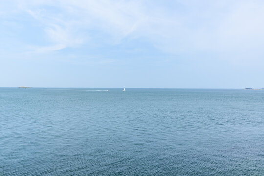 Serene seascape featuring calm blue water, a single sailboat and a speedboat on the horizon, and faint islands beneath a soft, lightly clouded sky.