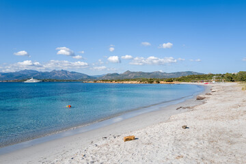 Fototapeta premium Bright white sand curves along the clear turquoise water at Spiaggia la Salinedda in Sardinia, with distant mountains and a yacht under a vivid blue sky dotted with small clouds.