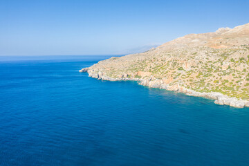 Obraz premium Wide aerial view of a rugged, sunlit rocky headland jutting into the deep blue Mediterranean waters near Preveli, Crete. The coastline features arid slopes with sparse greenery under a clear sky