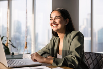 Obraz premium Happy successful Latin young business woman looking away from workplace, smiling, laughing, enjoying professional achievement, successful career. Candid portrait of businesswoman at table with laptop