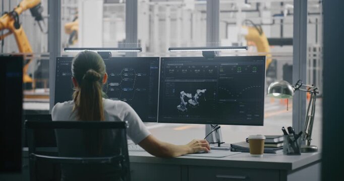 Focused Female Engineer Tracking Robotic Performance Metrics and Digital Twin Model on Workstation, With Factory in Background. Data Driven Decision Making and Real Time System Analysis. Back View.