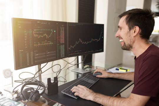 Happy successful investor man analyzing growths charts on two large monitors. Freelance trader working from home, managing assets on stock market exchange platform, getting profit