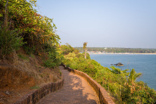 Curved stone walkway bordered by lush greenery overlooks the calm blue waters and sandy shoreline of Bekal, India. Soft sunlight enhances the natural textures and tranquil atmosphere.