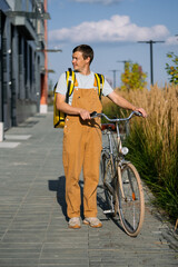 Male bike courier delivering food or parcel in city, walking with bicycle on sunny day, wearing overalls and bright yellow backpack, providing efficient urban delivery service