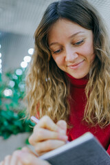 Young caucasian woman in a red turtleneck, curly hair, smiling as she writes in a small notebook with a pen, focused and content while journaling or planning ideas
