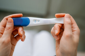 Woman holding white and blue pregnancy test in both hands. Studying single line negative result with tense. Intimate expression conveying uncertainty. Waiting. And reproductive health decision-making