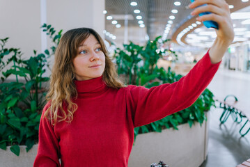 Young woman in a red turtleneck smiling while extending her arm, holding a smartphone to take a selfie in a large shopping mall with green plants and modern lighting in the background