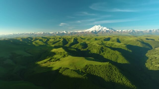 Aerial footage captures a stunning panoramic landscape of the majestic, snow-capped Mount Elbrus, Europe's highest mountain, situated in the Caucasus range. The foreground features vibrant green rolli