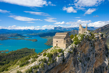 Fototapeta premium Ancient stone castle and church perch on a dramatic limestone ridge high above the vivid blue waters of Mediano lake in Aragon, Spain. Panoramic mountain views, lush greenery, and striking rock