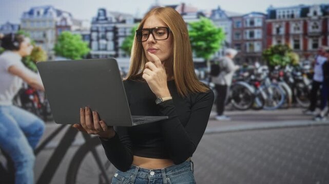Woman holding laptop touching chin with hand on a busy street beside rows of parked bicycles and historic buildings; thoughtful focus.