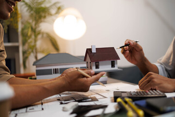 Two people are working on a house plan design. They are using various tools, working with the plan, and reviewing a house model to verify the accuracy of their work in the workplace.