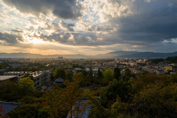 Fototapeta premium Sweeping view of Kyoto at sunset, with dramatic clouds, warm sunlight breaking through, and distant mountain silhouettes framing the city and lush autumn trees.
