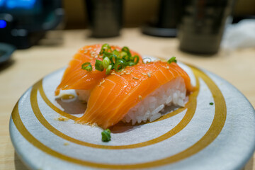 Two pieces of salmon nigiri sushi topped with chopped green onions and sesame seeds are served on a modern silver and gold plate. The fresh fish and glossy rice are highlighted by soft indoor lighting