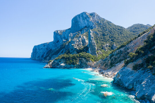 Jagged limestone cliffs tower above vibrant turquoise sea and lush green vegetation at Cala Goloritze, Baunei, Sardinia. Bright sunlight enhances the dramatic coastal landscape and crystal-clear water