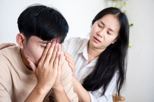 A worried young woman comforting a stressed Asian man covering his face in despair. Concept of mental health support, relationship problems, empathy, and emotional stress at home.