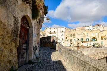 Fotobehang Smal steegje A narrow cobblestone path winds past an old stone building covered in graffiti, opening to sunlit historic facades in Matera, Italy under a bright sky with scattered clouds.  © Florent