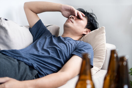Exhausted Asian man lying on a sofa feeling sick with a hangover after drinking beer. Young male suffering from a headache, dehydration, and alcoholism. Health problems and substance abuse concept.