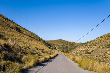 A narrow paved road stretches between sunlit grassy slopes and rugged hills at Cap Tenare, Greece....
