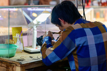 A jewelry artisan in a colorful plaid shirt works intently at a cluttered wooden workbench under a bright lamp in Battambang market, Cambodia. The workspace features scattered tools, a green plastic