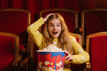 Surprised child girl watching movie in cinema, holding popcorn bucket, sitting in red theater seats with excited facial expression.