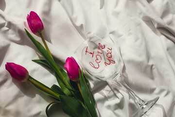 I LOVE YOU message on wine glass with fresh tulips on white bed with trendy deep shadows. Spring flowers in pink violet colors. Romantic postcard wallpaper design Copy space 