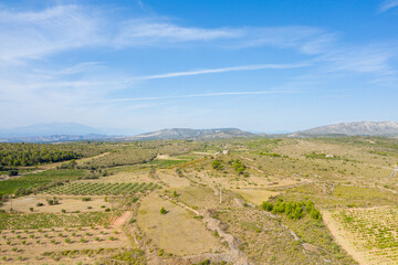 Wide aerial view of sunlit fields, vineyards, and scattered trees stretching toward distant hills and mountains near Opoul, with a clear sky and soft clouds above.
