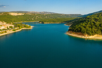 Fototapeta premium Aerial view of the vivid blue waters of Lac de Sainte-Croix curving between forested hills and the sunlit village of Bauduen, with gentle slopes and clear summer skies.