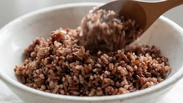 A close-up shot elegantly captures the simple beauty of cooked whole grains being served. A rustic wooden spoon gently scoops a portion of the rich, reddish-brown grains from a white ceramic bowl, hig