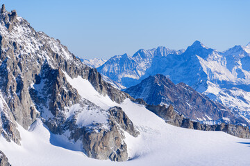 Steep, rugged cliffs dusted with snow rise above the sweeping expanse of Glacier du Geant, with layers of jagged blue mountains fading into the distance under a clear sky.